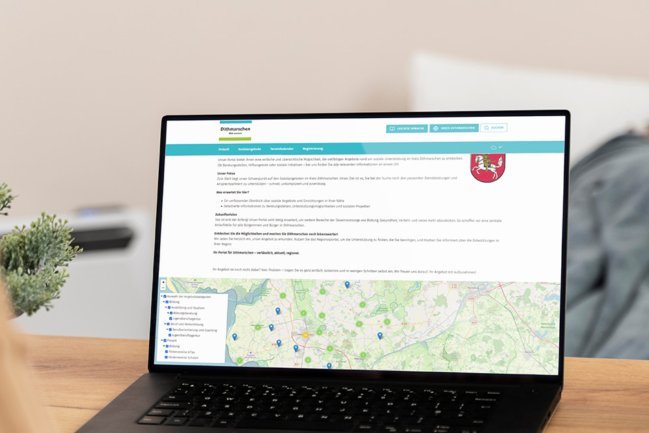 Eine Frau betrachtet das neue Regionalportal Dithmarschen auf dem Monitor und sucht nach Gesundheitsangeboten. Im Hintergrund liegt ein kranker Mann im Bett.